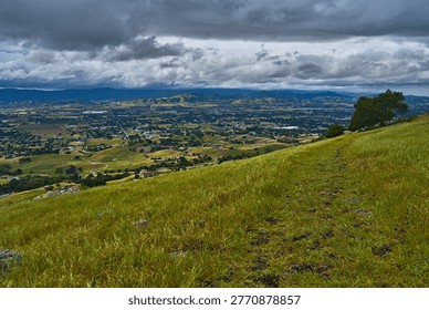A dramatic panoramic view from a grassy hillside overlooking a vast green valley with patchwork fields, a sprawling town, and distant mountains under a moody, cloudy sky.