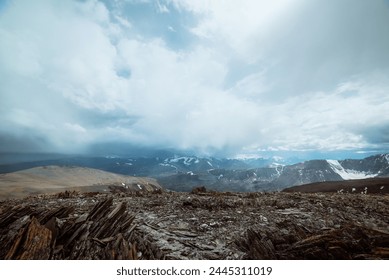Dramatic panoramic view from high stony pass to alpine valley against large snow-capped mountain range in rainy low clouds. Sharp stones on hill against misty snowy mountains in rain under cloudy sky.