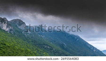 A dramatic panoramic view of the mountains in northern Corfu, Greece. The green slopes and rocky peaks are shrouded in low, heavy fog and dark, moody storm clouds.