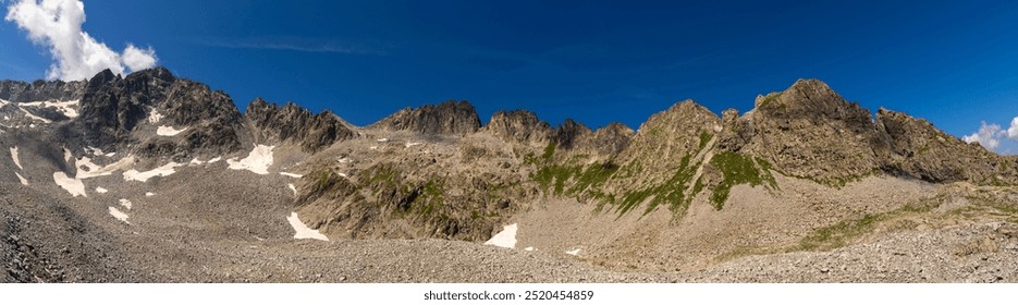 A dramatic panoramic view of rugged mountain peaks with patches of snow and expansive rocky terrain, set against a deep blue sky, showcasing the raw beauty of the highlands