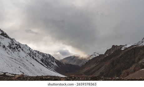 Dramatic panoramic view of snow-capped mountains under a cloudy sky.  The rugged terrain and the interplay of light and shadow create a breathtaking scene.  A powerful image of nature's grandeur.