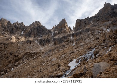 A dramatic panoramic view of a vast, rugged mountain range featuring sharp rocky peaks, steep slopes covered in scree, and scattered patches of snow under a cloudy sky.