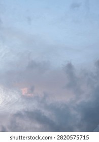 Dramatic and peaceful sky background during sunset or dawn, featuring dark grey cloudscape mixed with soft pastel pink and orange sunlight on a light blue atmosphere.