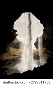 A dramatic perspective from behind the cascading curtain of water at Kvernufoss, a hidden gem waterfall in South Iceland, showcasing the unique experience of walking behind the falls through a natural rock formation, with the misty landscape and gorge visible through the waterfall’s frame.