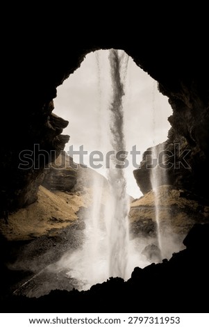 A dramatic perspective from behind the cascading curtain of water at Kvernufoss, a hidden gem waterfall in South Iceland, showcasing the unique experience of walking behind the falls through a natural rock formation, with the misty landscape and gorge visible through the waterfall’s frame.
