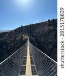 Dramatic perspective looking down a narrow suspension bridge spanning a deep mountain gorge in Kyrgyzstan. Metal mesh railings create strong leading lines converging at the far end where the bridge meets a rocky cliff with conifer trees. Bright sun in clear blue sky overhead. Adventure travel concept.