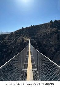 Dramatic perspective looking down a narrow suspension bridge spanning a deep mountain gorge in Kyrgyzstan. Metal mesh railings create strong leading lines converging at the far end where the bridge meets a rocky cliff with conifer trees. Bright sun in clear blue sky overhead. Adventure travel concept.
