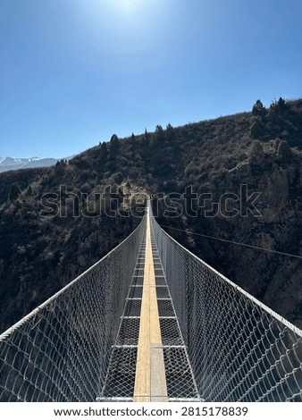 Dramatic perspective looking down a narrow suspension bridge spanning a deep mountain gorge in Kyrgyzstan. Metal mesh railings create strong leading lines converging at the far end where the bridge meets a rocky cliff with conifer trees. Bright sun in clear blue sky overhead. Adventure travel concept.