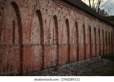 A dramatic perspective shot looking down the length of a long, historic red brick wall. The structure is defined by a rhythmic series of tall, arched decorative niches. The weathered bricks show a rich texture of age, standing above a traditional cobblestone base under the soft, muted light of an overcast evening.