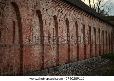 A dramatic perspective shot looking down the length of a long, historic red brick wall. The structure is defined by a rhythmic series of tall, arched decorative niches. The weathered bricks show a rich texture of age, standing above a traditional cobblestone base under the soft, muted light of an overcast evening.