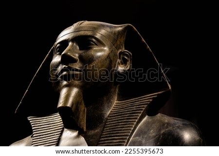 Dramatic photo of the face of a statue of Ramses in an Egyptian museum with a dark background. The details and expression of the pharaoh stand out against the contrast of light and shadow.