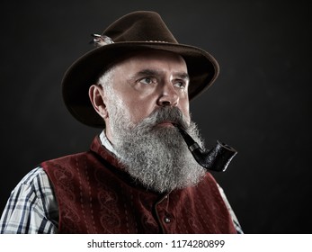 Dramatic Portrait Of Gray Bearded Senior Man In Hat Smoking Tobacco Pipe. View Of Austrian, Tyrolean, Bavarian Old Man In National Traditional Costume In Retro Style.