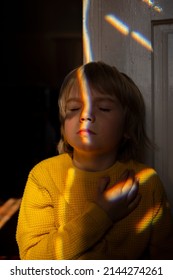 Dramatic Portrait Of A Lonely Ukrainian Boy 6 Years Old In A Yellow Sweater In A Dark Room. Sun Iridescent Glare On The Face, Light Refraction, Distortion. A Ray Of Light As A Symbol Of Hope And Joy