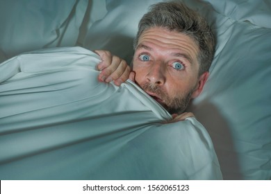 Dramatic Portrait Of Stressed And Scared Man Alone In Bed Awake At Night In Fear After Having A Nightmare Feeling Paranoid Holding The Blanket In Funny Panic Face Expression 
