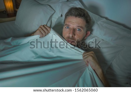 dramatic portrait of stressed and scared man alone in bed awake at night in fear after having a nightmare feeling paranoid holding the blanket in funny panic face expression 
