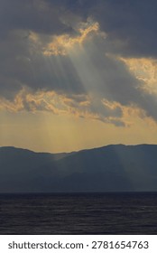 Dramatic rays of sunlight breaking through the clouds over the mountains. Divine or twilight rays breaking through thick clouds onto a dark mountain range and the sea below.
