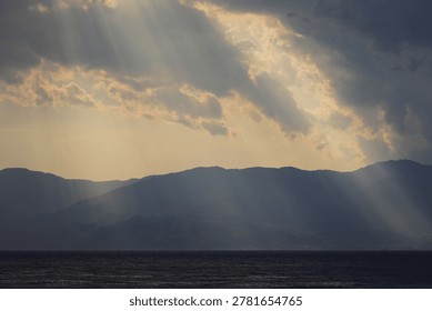 Dramatic rays of sunlight breaking through the clouds over the mountains. Divine or twilight rays breaking through thick clouds onto a dark mountain range and the sea below.