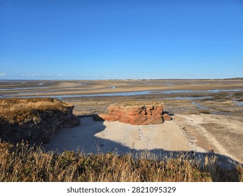 Dramatic red sandstone cliffs and exposed rock bed along the coastline of Hilbre Island at low tide under a bright blue sky, weathered red sandstone rock formations displaying natural erosion layers on the shore 