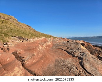 Dramatic red sandstone cliffs and exposed rock bed along the coastline of Hilbre Island at low tide under a bright blue sky, weathered red sandstone rock formations displaying natural erosion layers on the shore 