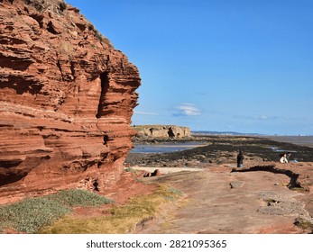 Dramatic red sandstone cliffs and exposed rock bed along the coastline of Hilbre Island at low tide under a bright blue sky, weathered red sandstone rock formations displaying natural erosion layers on the shore 