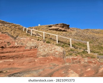 Dramatic red sandstone cliffs and exposed rock bed along the coastline of Hilbre Island at low tide under a bright blue sky, weathered red sandstone rock formations displaying natural erosion layers on the shore 