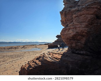 Dramatic red sandstone cliffs and exposed rock bed along the coastline of Hilbre Island at low tide under a bright blue sky, weathered red sandstone rock formations displaying natural erosion layers on the shore