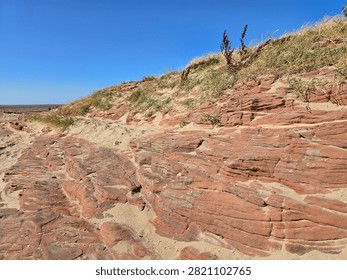Dramatic red sandstone cliffs and exposed rock bed along the coastline of Hilbre Island at low tide under a bright blue sky, weathered red sandstone rock formations displaying natural erosion layers on the shore