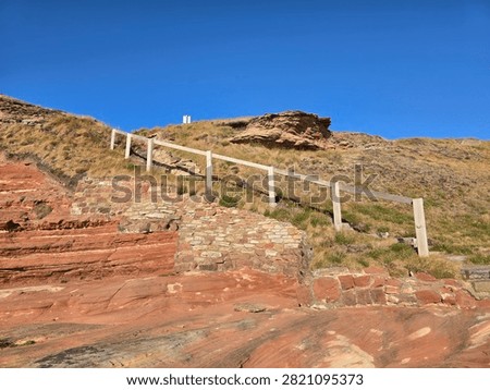 Dramatic red sandstone cliffs and exposed rock bed along the coastline of Hilbre Island at low tide under a bright blue sky, weathered red sandstone rock formations displaying natural erosion layers on the shore 