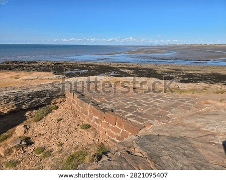 Dramatic red sandstone cliffs and exposed rock bed along the coastline of Hilbre Island at low tide under a bright blue sky, weathered red sandstone rock formations displaying natural erosion layers on the shore 