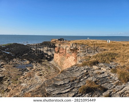 Dramatic red sandstone cliffs and exposed rock bed along the coastline of Hilbre Island at low tide under a bright blue sky, weathered red sandstone rock formations displaying natural erosion layers on the shore 
