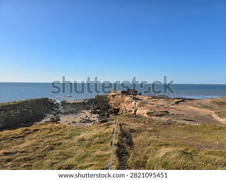 Dramatic red sandstone cliffs and exposed rock bed along the coastline of Hilbre Island at low tide under a bright blue sky, weathered red sandstone rock formations displaying natural erosion layers on the shore 