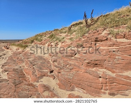 Dramatic red sandstone cliffs and exposed rock bed along the coastline of Hilbre Island at low tide under a bright blue sky, weathered red sandstone rock formations displaying natural erosion layers on the shore