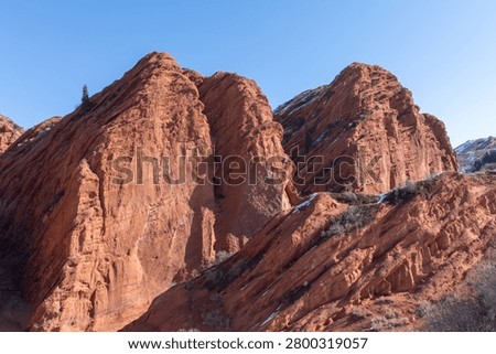 Dramatic red sandstone cliffs of the Seven Bulls rock formation in Jeti-Oguz Gorge, Issyk-Kul Region, Kyrgyzstan, with patches of snow under a clear blue sky