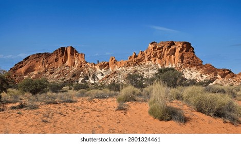 Dramatic red sandstone rock formation rising above sandy scrubland and low bushes, set against a vivid blue sky. Rainbow Valley Northern Territory in Australia.