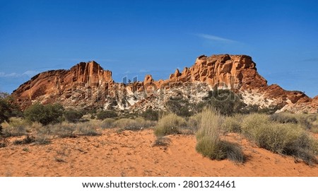 Dramatic red sandstone rock formation rising above sandy scrubland and low bushes, set against a vivid blue sky. Rainbow Valley Northern Territory in Australia.