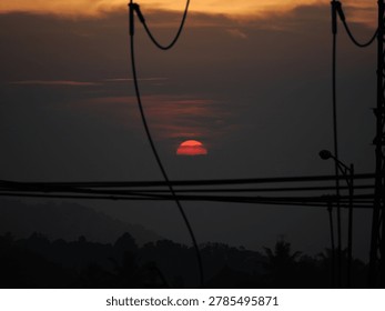 Dramatic red sun setting behind dark clouds and silhouette of street power lines over a distant misty hill in the evening