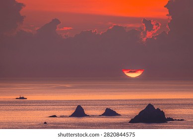 Dramatic red sunrise beneath dark clouds over the Pacific Ocean with a fishing boat and rocky coastline in Yilan, Taiwan.