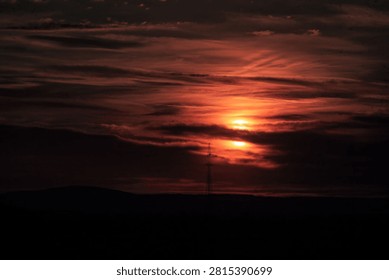 Dramatic red sunset behind dark clouds over a silhouetted horizon with a lone tower, creating a moody evening sky