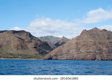 Dramatic reddish layered volcanic sea cliffs with deep ravines rise steeply from the deep blue Atlantic on the rugged coast of La Gomera under a partly cloudy sky.