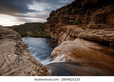 A dramatic river canyon cuts through rugged rocky terrain with towering sandstone cliffs and flowing water, captured during golden hour with moody cloud cover overhead.