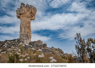 Dramatic rock formation resembling a stone pillar rising from a rugged landscape, with a lone hiker standing on rocky terrain beneath a cloudy sky. Concept of scale, exploration, adventure, solitude, and the power of nature in a remote wilderness setting.