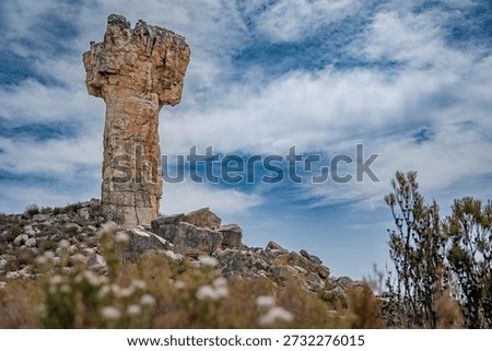 Dramatic rock formation resembling a stone pillar rising from a rugged landscape, with a lone hiker standing on rocky terrain beneath a cloudy sky. Concept of scale, exploration, adventure, solitude, and the power of nature in a remote wilderness setting.