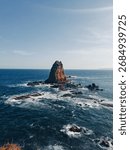 Dramatic rock formation rises from the dark blue ocean, surrounded by waves, under a clear sky, creating a tranquil and powerful scene ideal for travel and nature-themed publications.