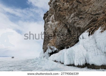 Dramatic rocky cliff covered with icicles and blue ice on the frozen shore of Lake Baikal, Siberia, Russia, under cloudy sky with vast winter lake panorama