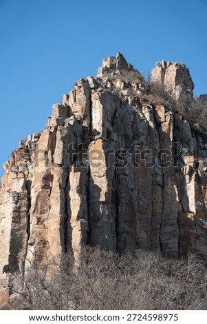 Dramatic rocky cliff rises against a clear blue sky in Armenia, vertical stone formations, warm earth tones, rugged mountain landscape