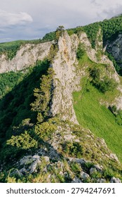Dramatic rocky cliff surrounded by lush forest and green hills under a bright summer sky, captured in daylight.