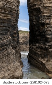 Dramatic rocky cliffs forming a narrow passage with ocean water below, showcasing rugged coastal geology and a glimpse of blue sky
