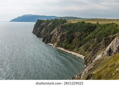 Dramatic rocky cliffs lined with dense Siberian taiga forest plunge toward the deep green waters of Lake Baikal along the eastern shoreline of Olkhon Island, with a distant mountain ridge visible across the water. Soft, diffused overcast light eliminates harsh shadows and reveals the full tonal range of the ochre and grey rock faces, emerald forest canopy, and the lake's characteristic blue-green hue. Available for editorial and commercial licensing.