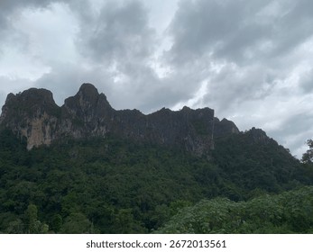 A dramatic rocky mountain range covered with green forest under a cloudy sky, natural outdoor landscape background.