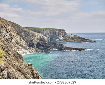 Dramatic rocky sea cliffs overlook clear turquoise water and a calm blue ocean under a bright cloudy sky in a wide scenic coastal landscape.